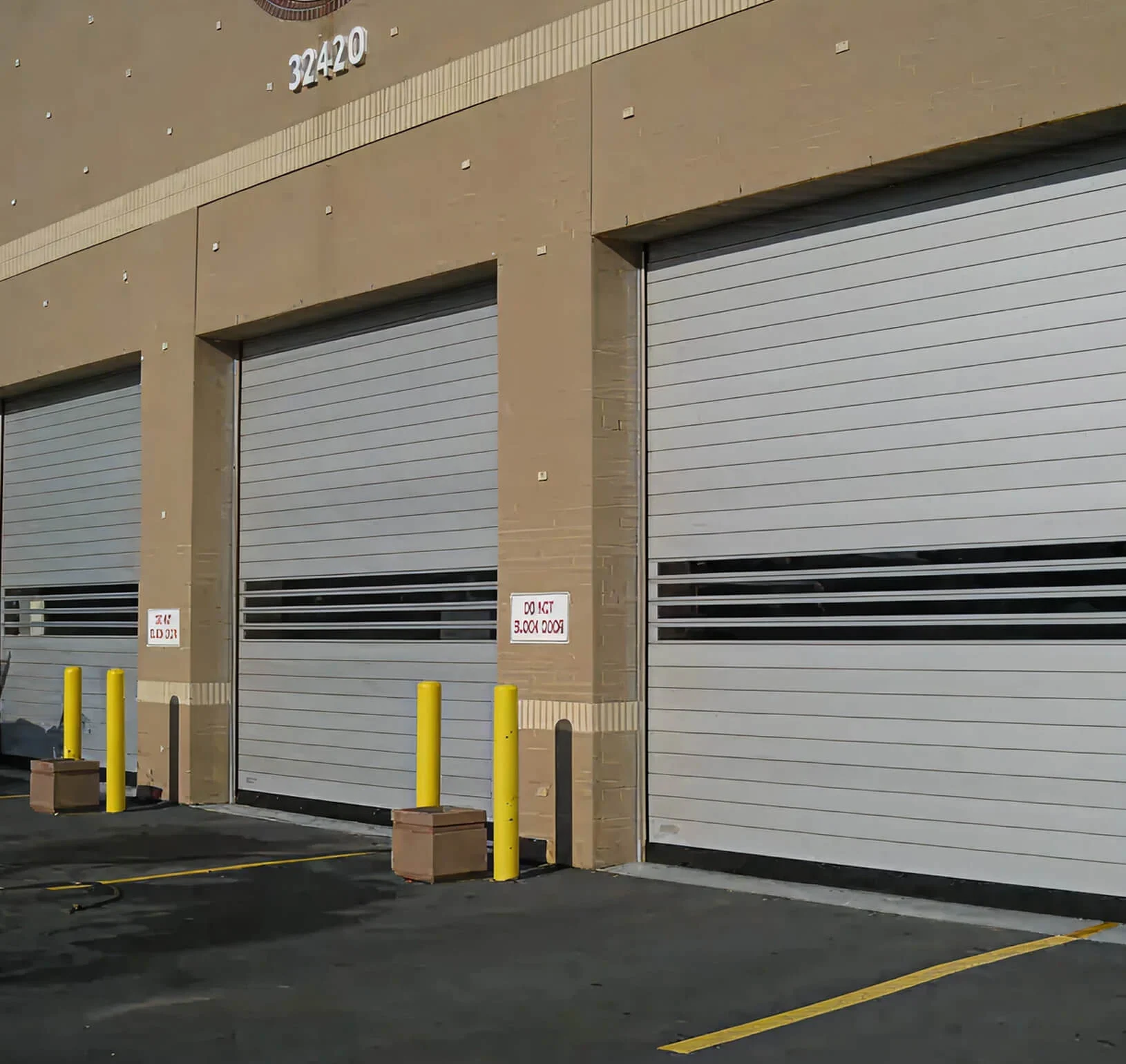 Three grey high-speed rigid rolling doors with vision slats on a tan commercial building, yellow bollards in front