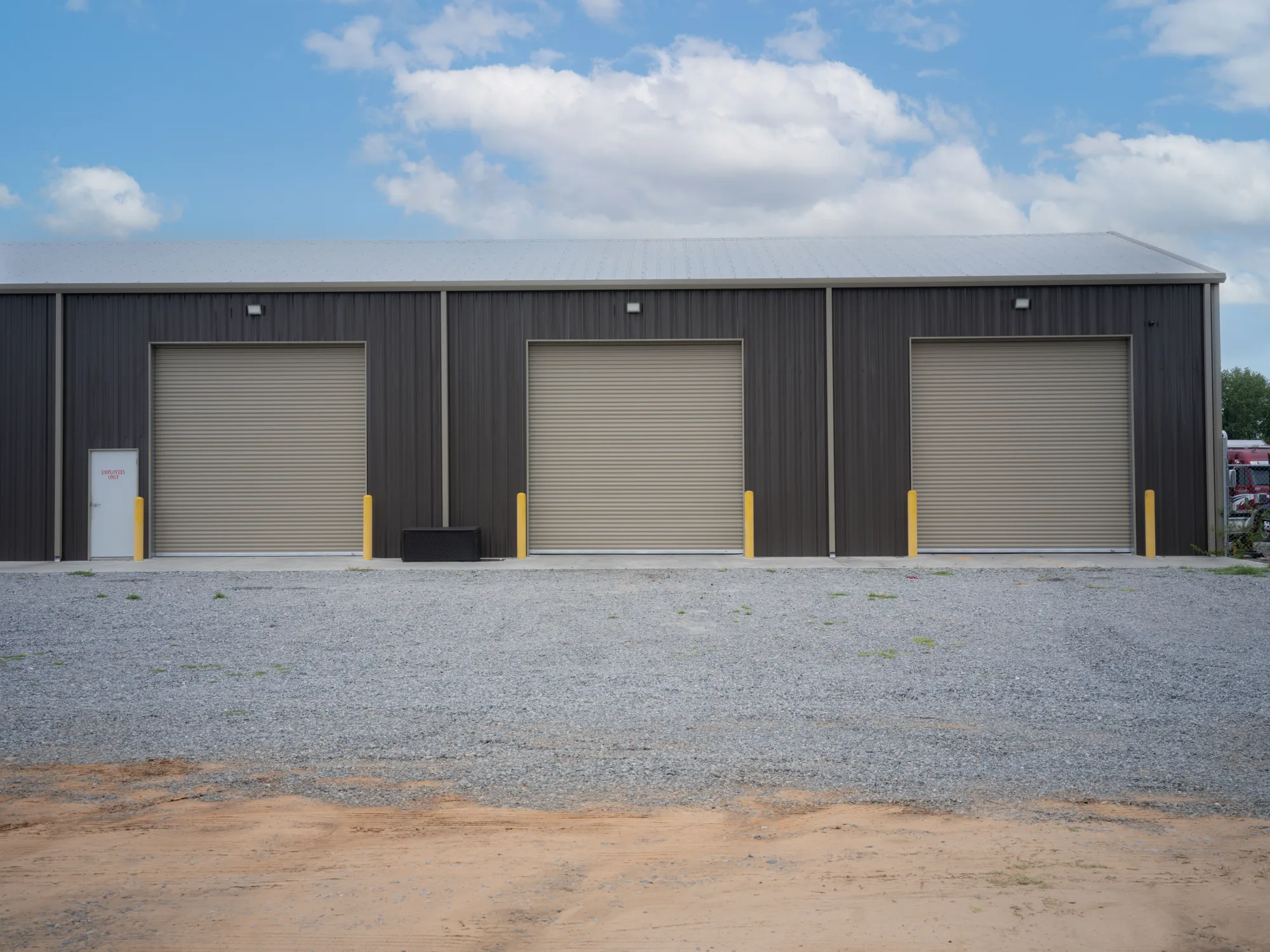 Three tan roll-up steel doors with yellow bollards on a dark grey metal shop building