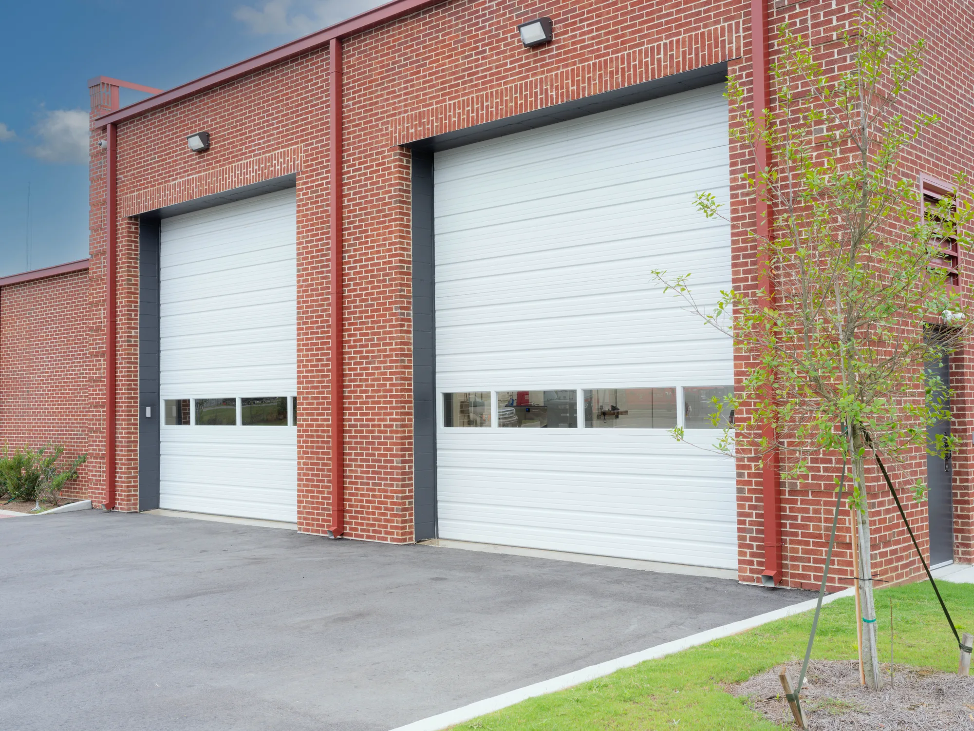 Two tall white sectional steel doors with a row of windows, set in a red brick building with red downspouts