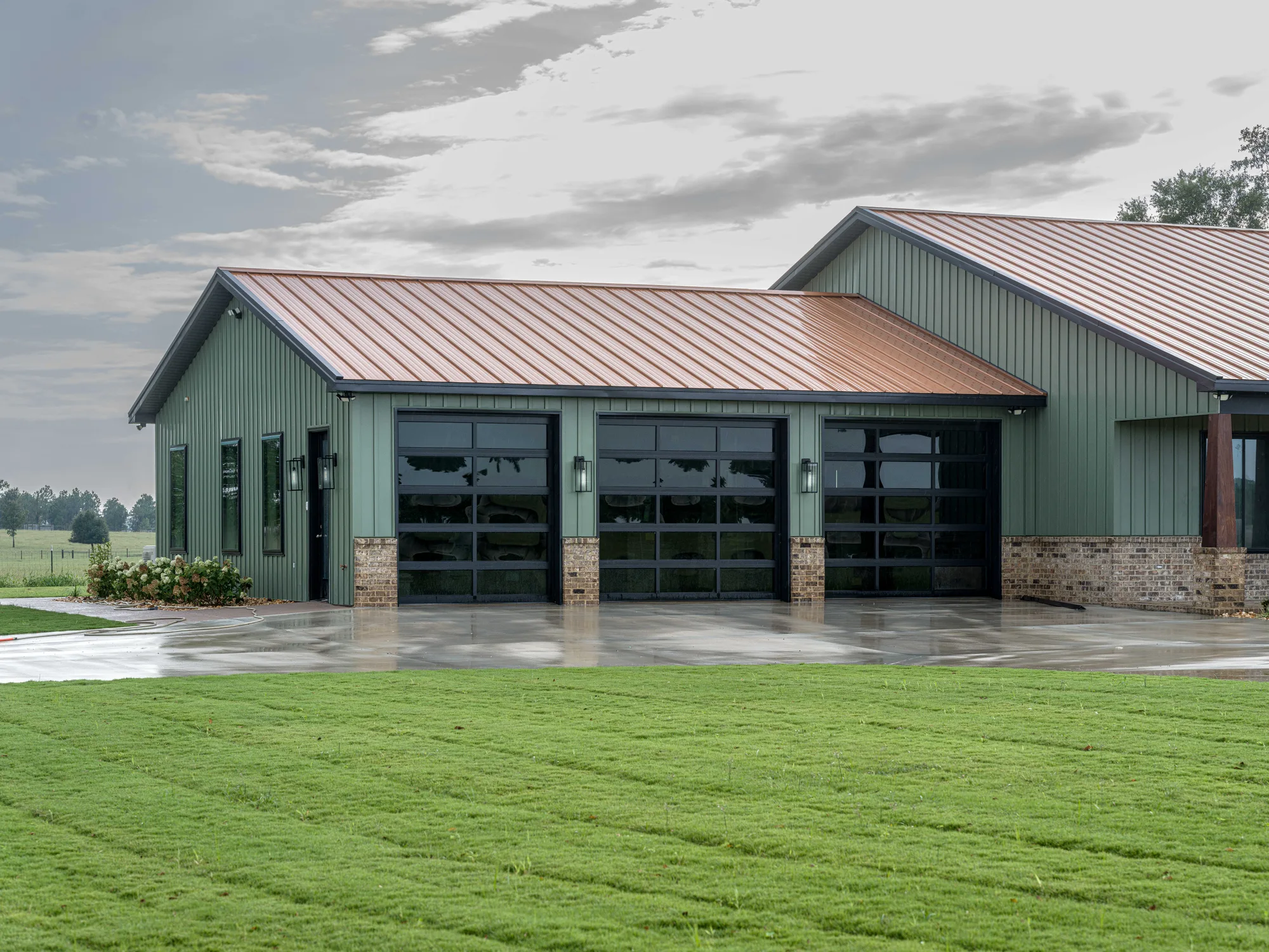 Three black-framed full-view glass garage doors on a sage green metal home with a copper-colored roof