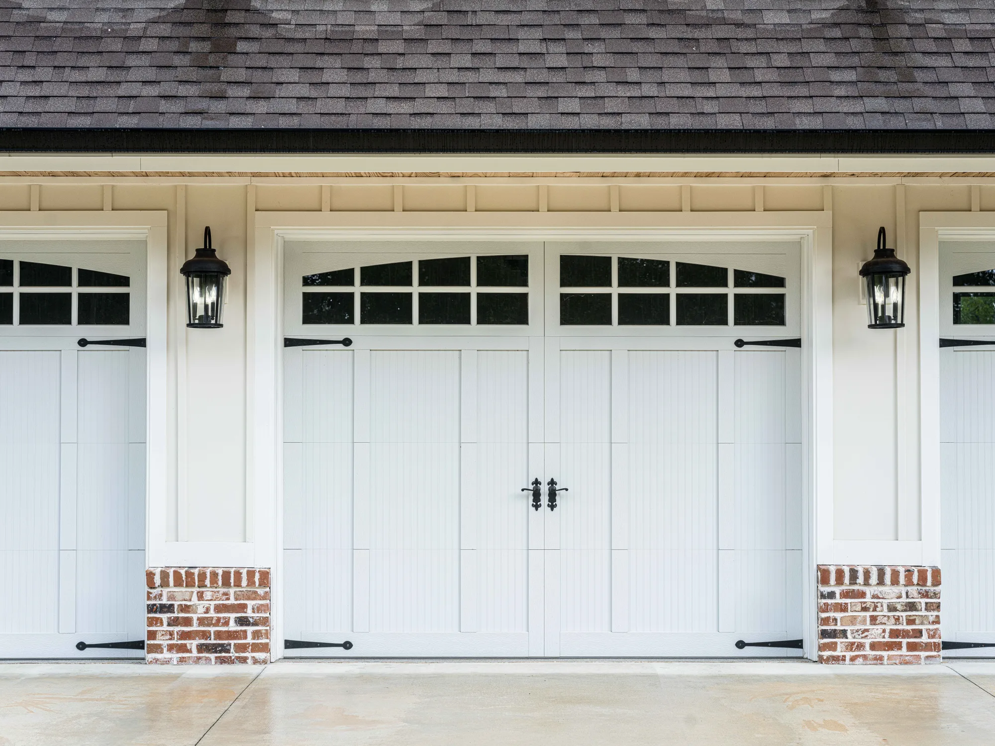 White carriage-style garage door with arched top windows, black strap hinges and handles, between brick-footed posts