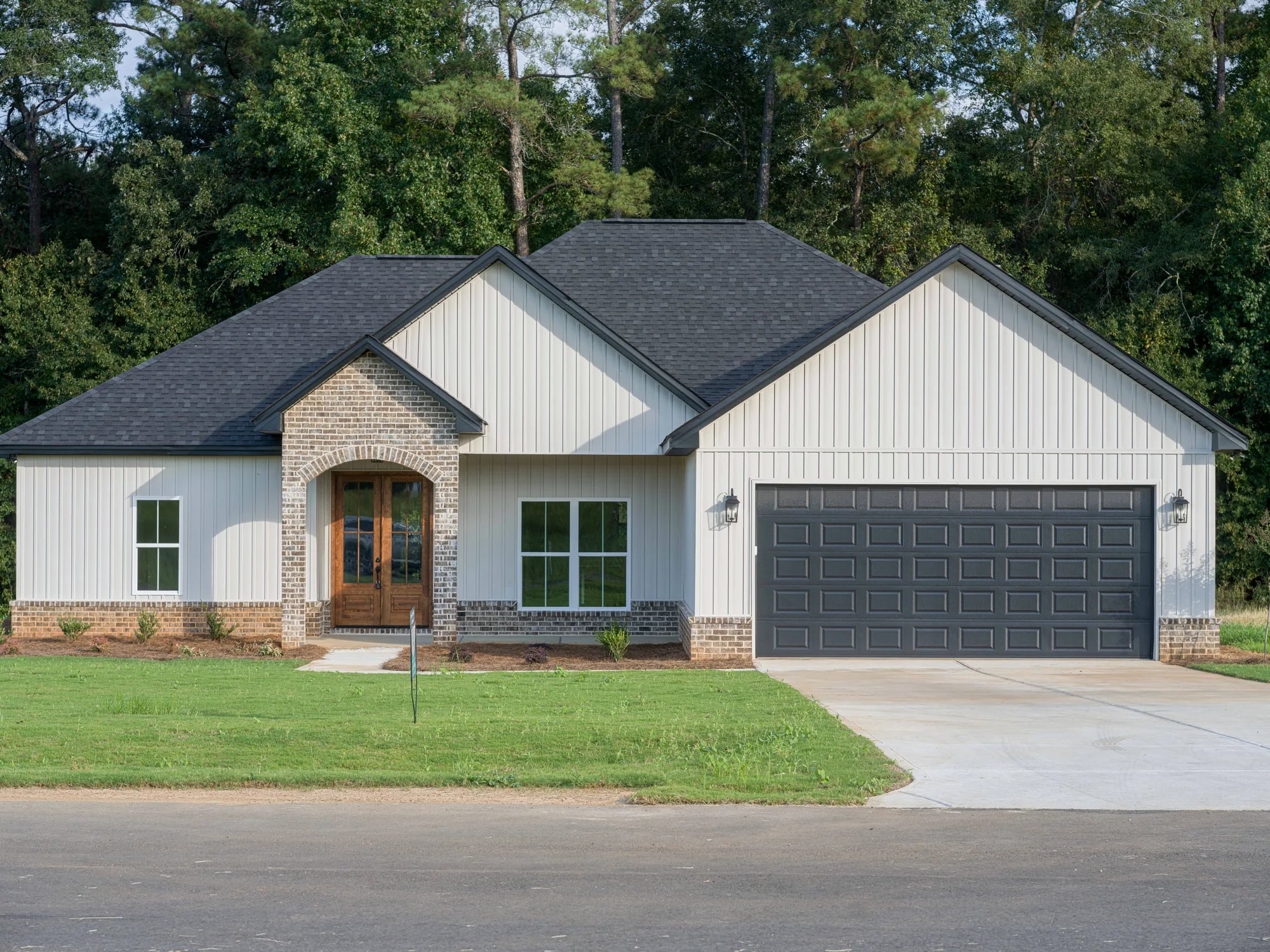 Off-white board-and-batten home with a brick entry arch and a charcoal raised-panel double garage door, seen from the street