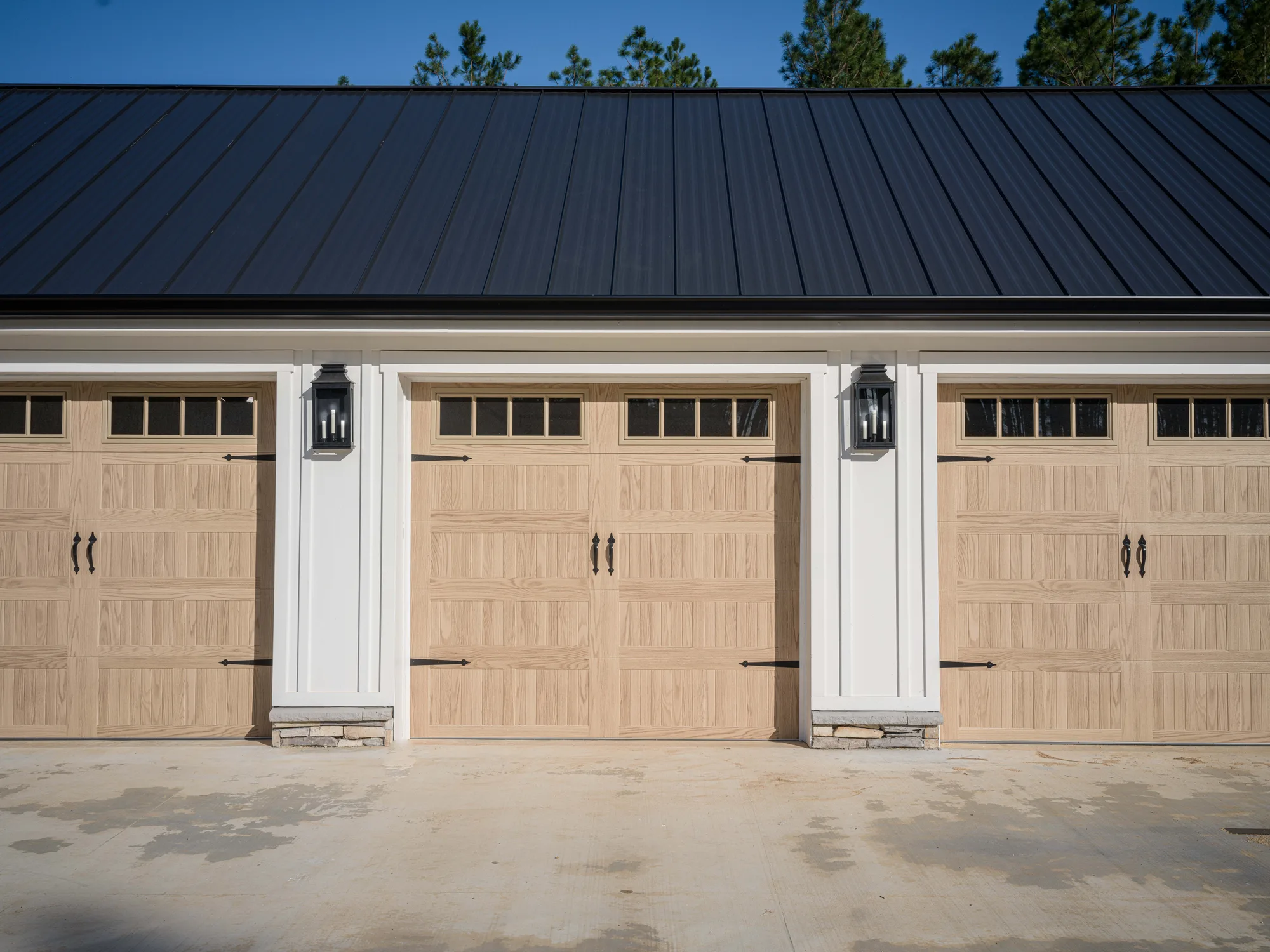 Light wood-look carriage-style garage doors with square top windows and black hardware on a white home with a black metal roof