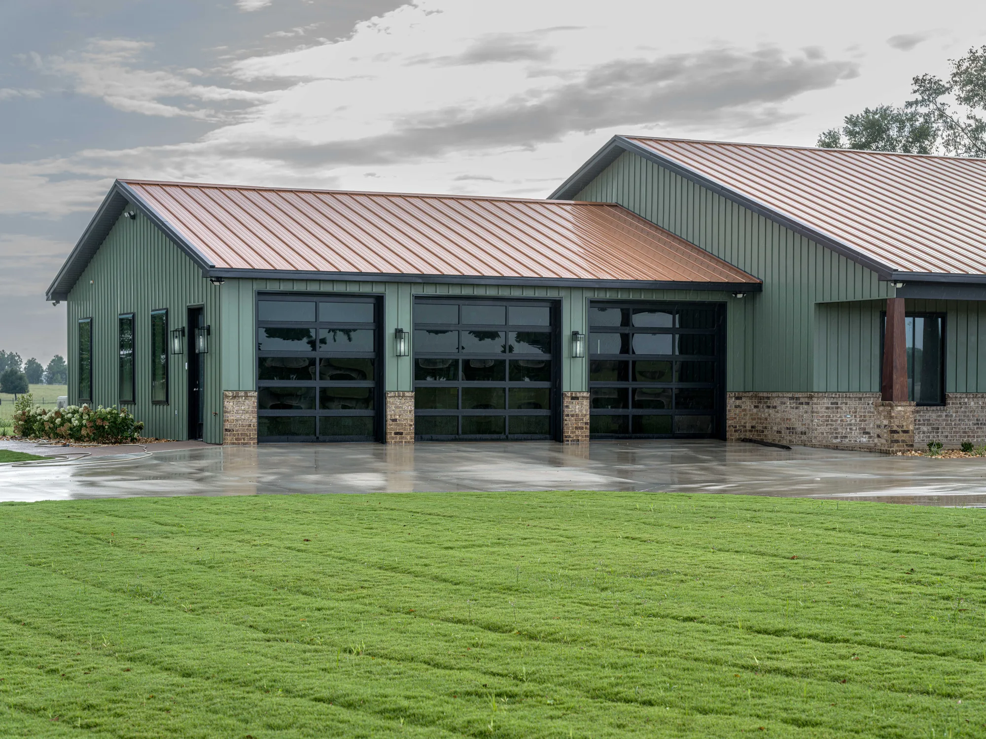 Three black-framed full-view glass garage doors on a sage green metal home with a copper-colored roof