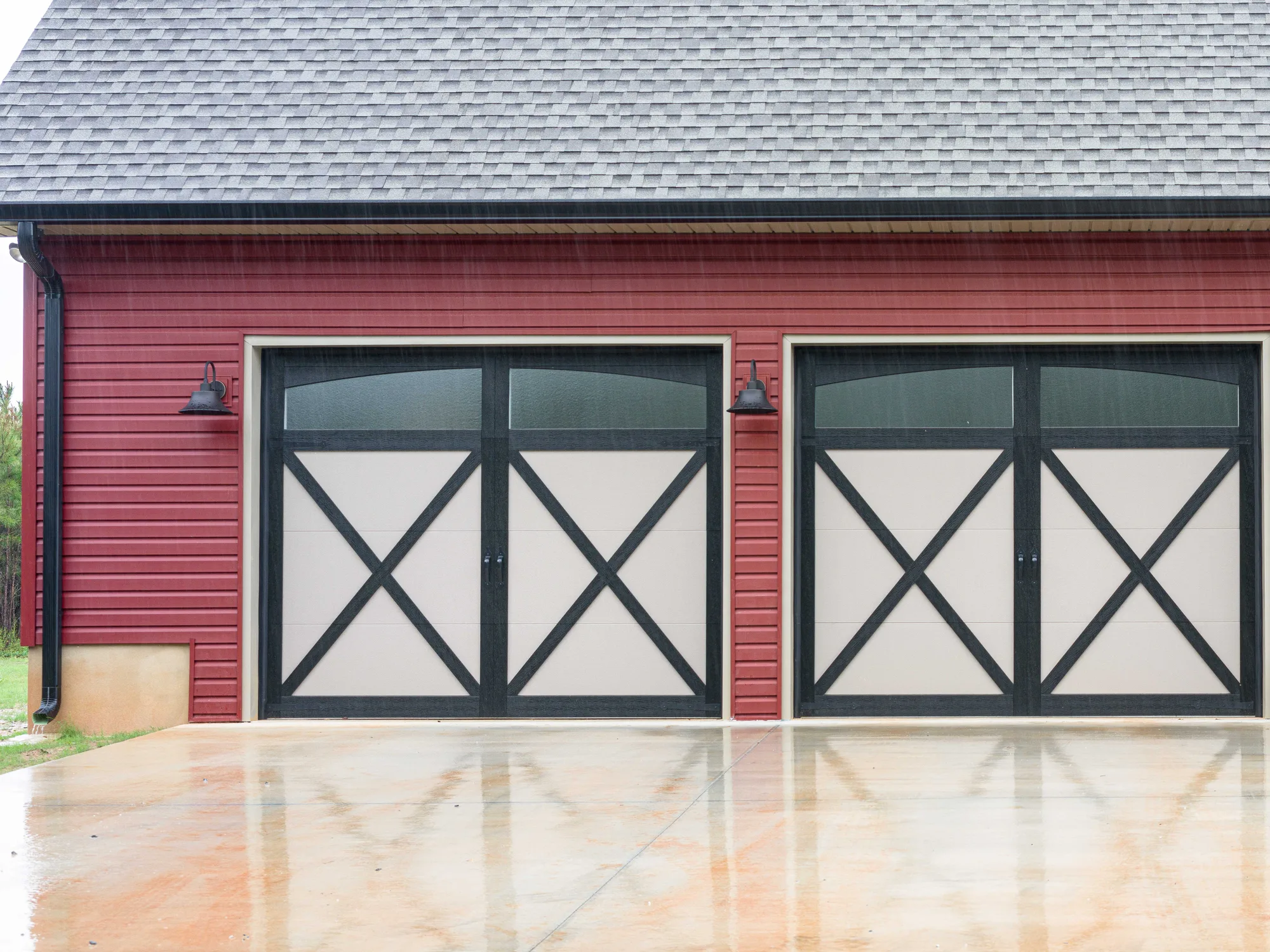 Two black and tan overlay garage doors with X-bracing and arched top glass on a red garage with a grey shingle roof