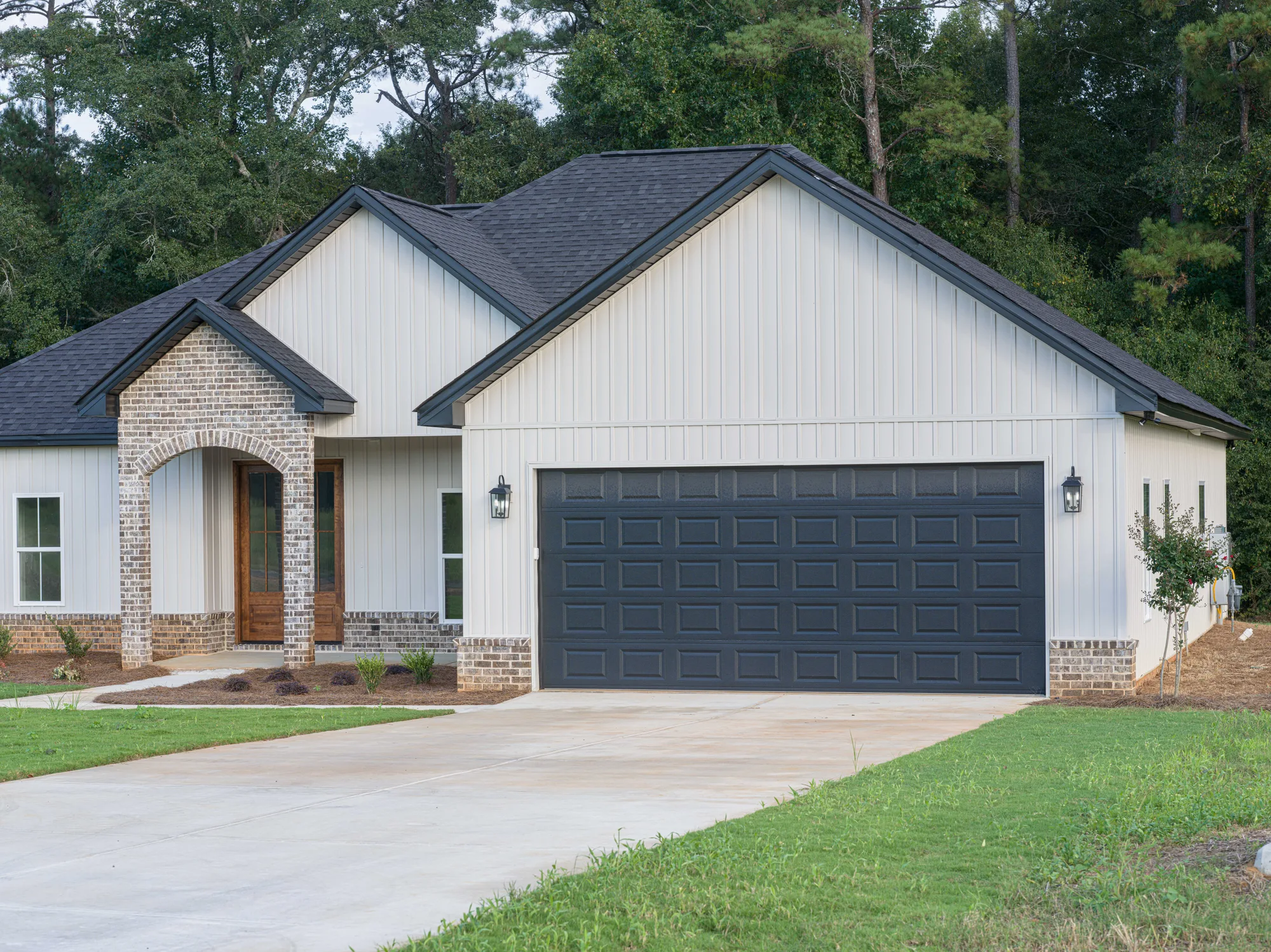 Charcoal raised-panel double garage door on an off-white board-and-batten home with a brick entry arch