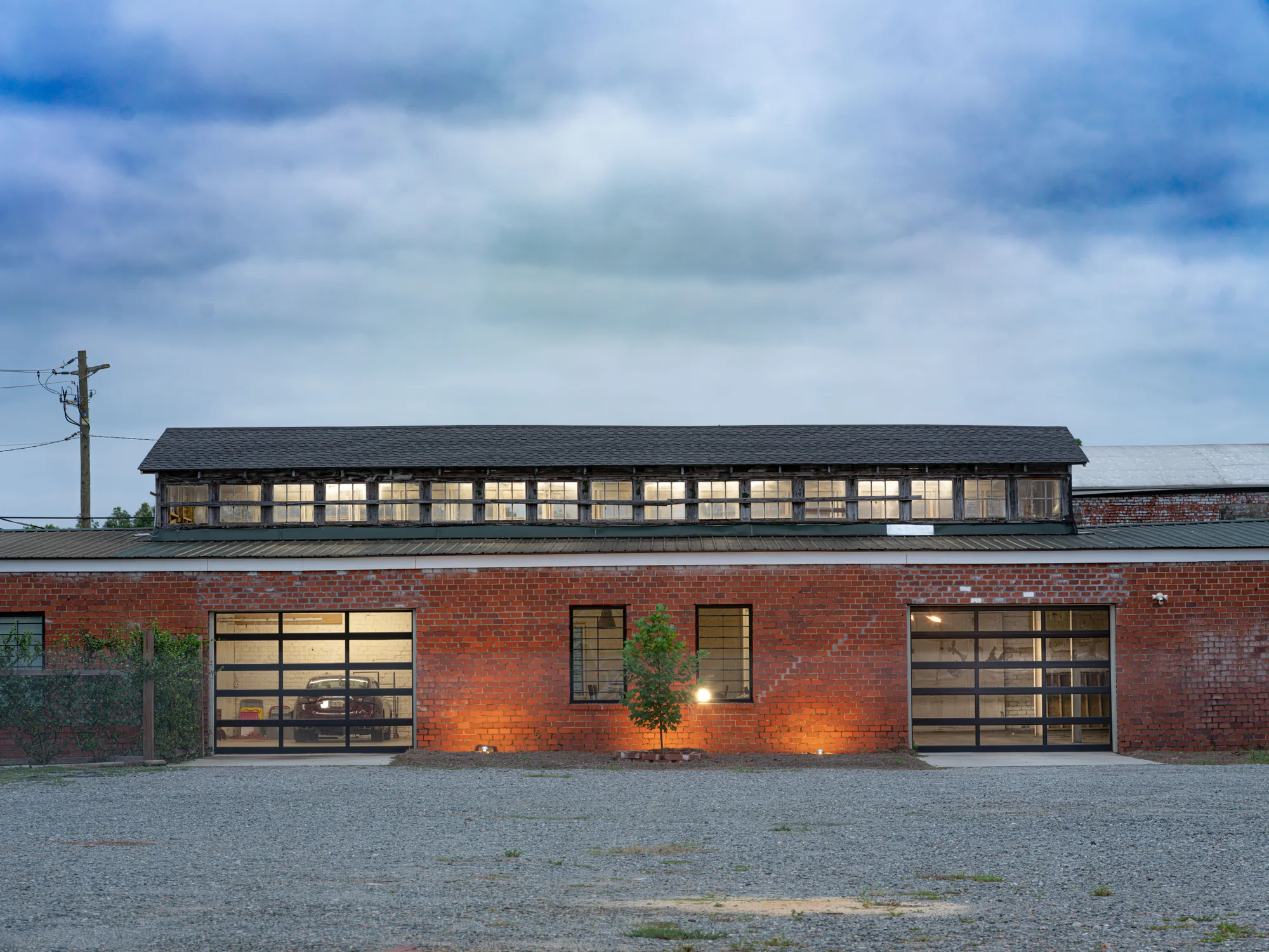 Two black-framed full-view glass doors on a long brick workshop with a lit clerestory at dusk