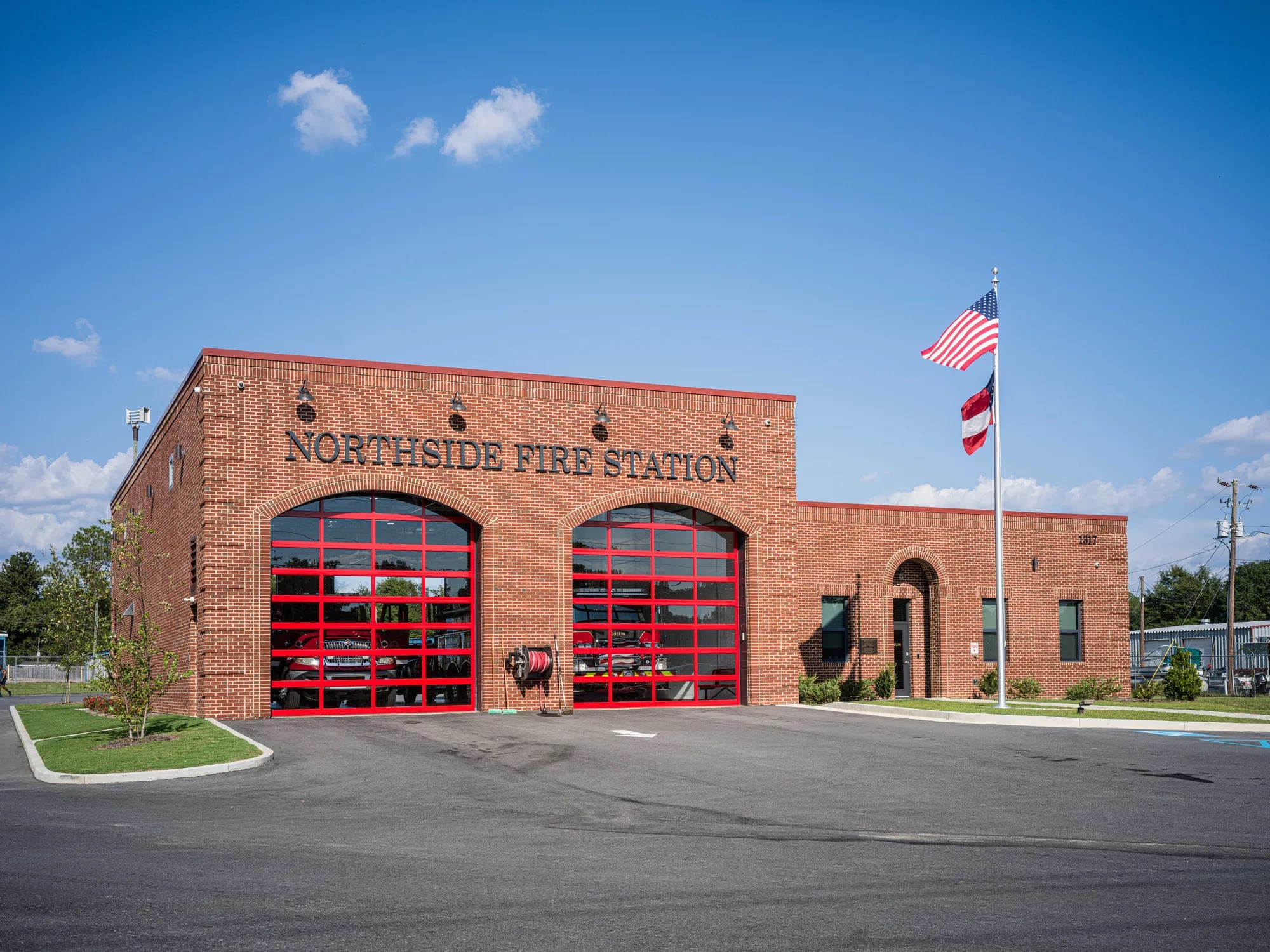 Two red-framed full-view glass doors in arched bays on a red brick fire station, with fire engines visible through the glass
