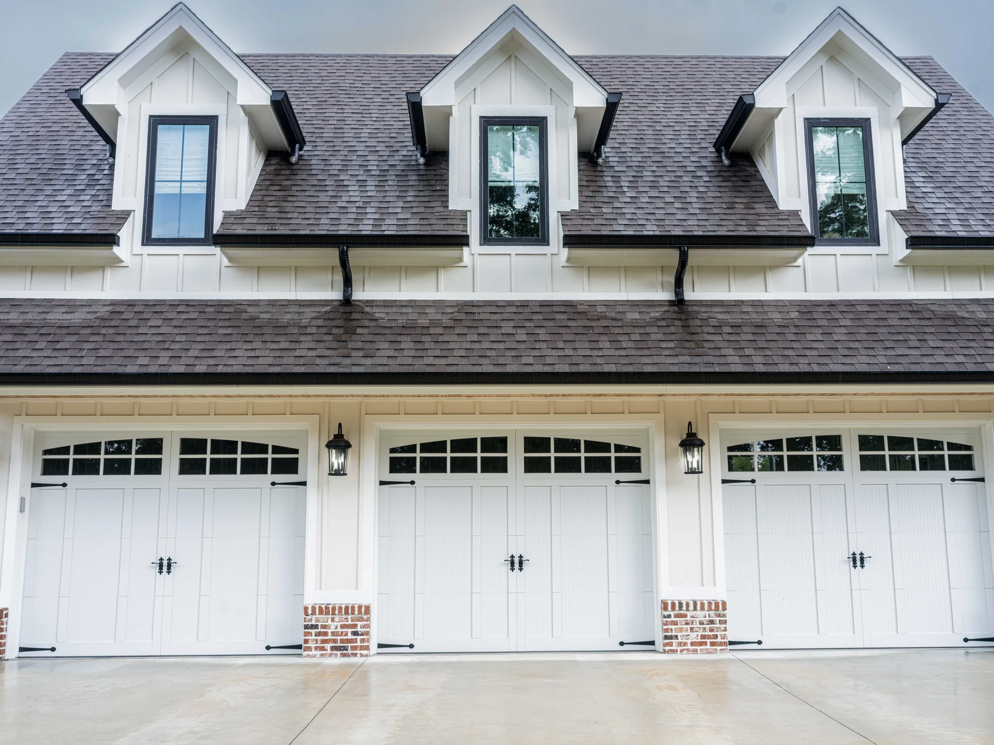 Three white carriage-style garage doors with arched top windows and black hardware under three dormers on a white board-and-batten garage