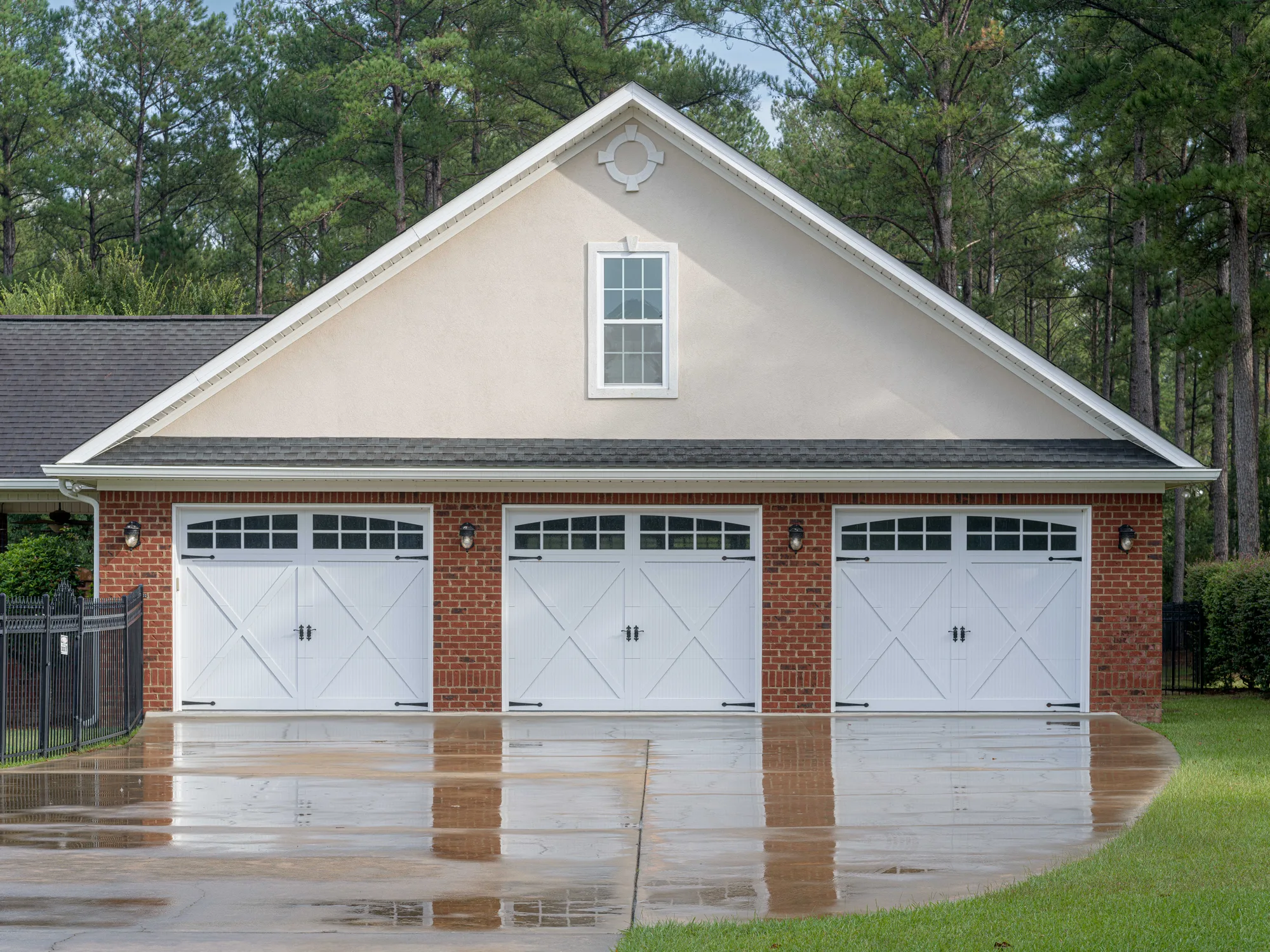 Three white crossbuck carriage-style garage doors with arched top windows on a red brick garage with a stucco gable