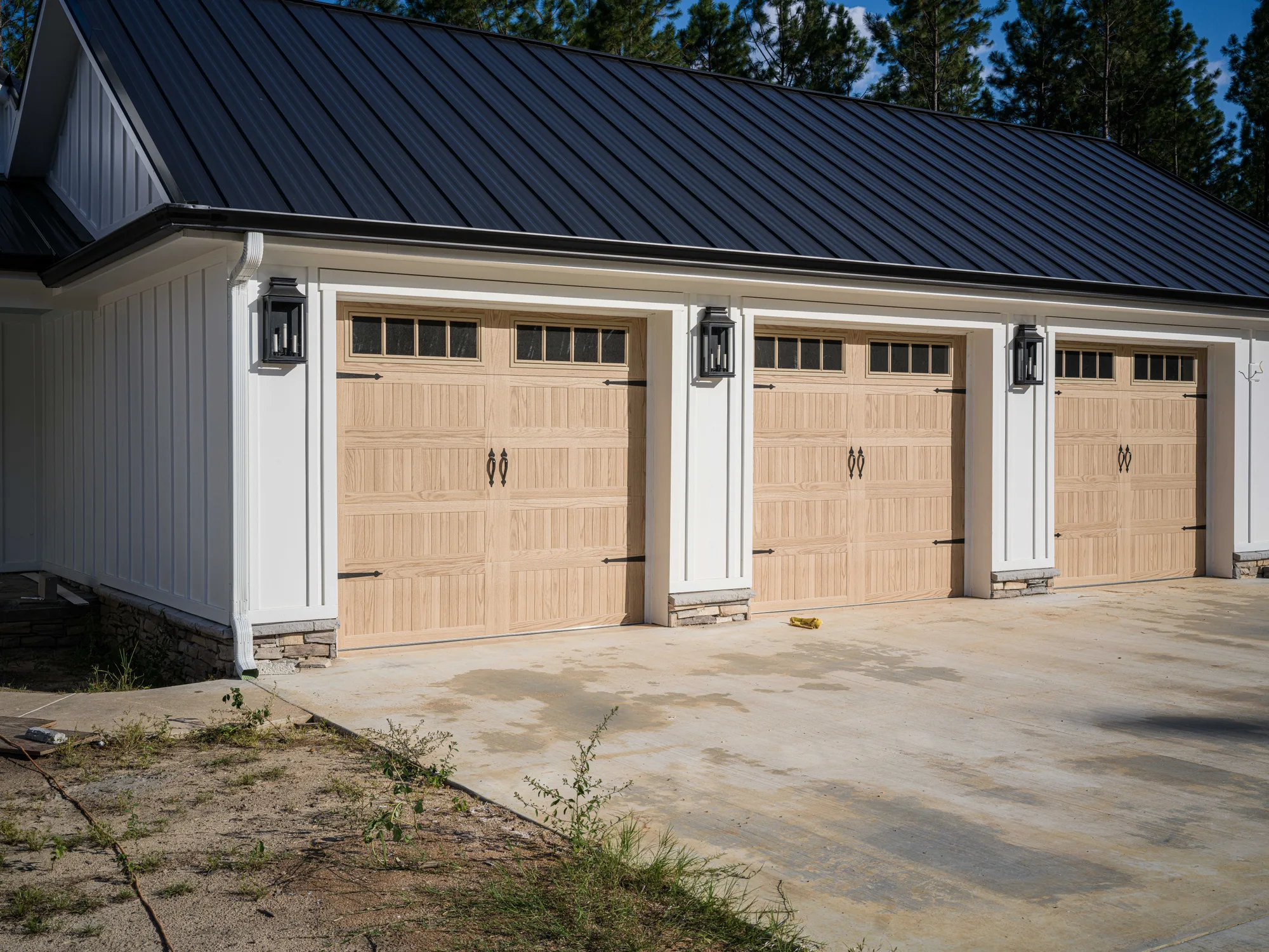 Three light wood-look carriage-style garage doors with square top windows on a white board-and-batten farmhouse with a black metal roof