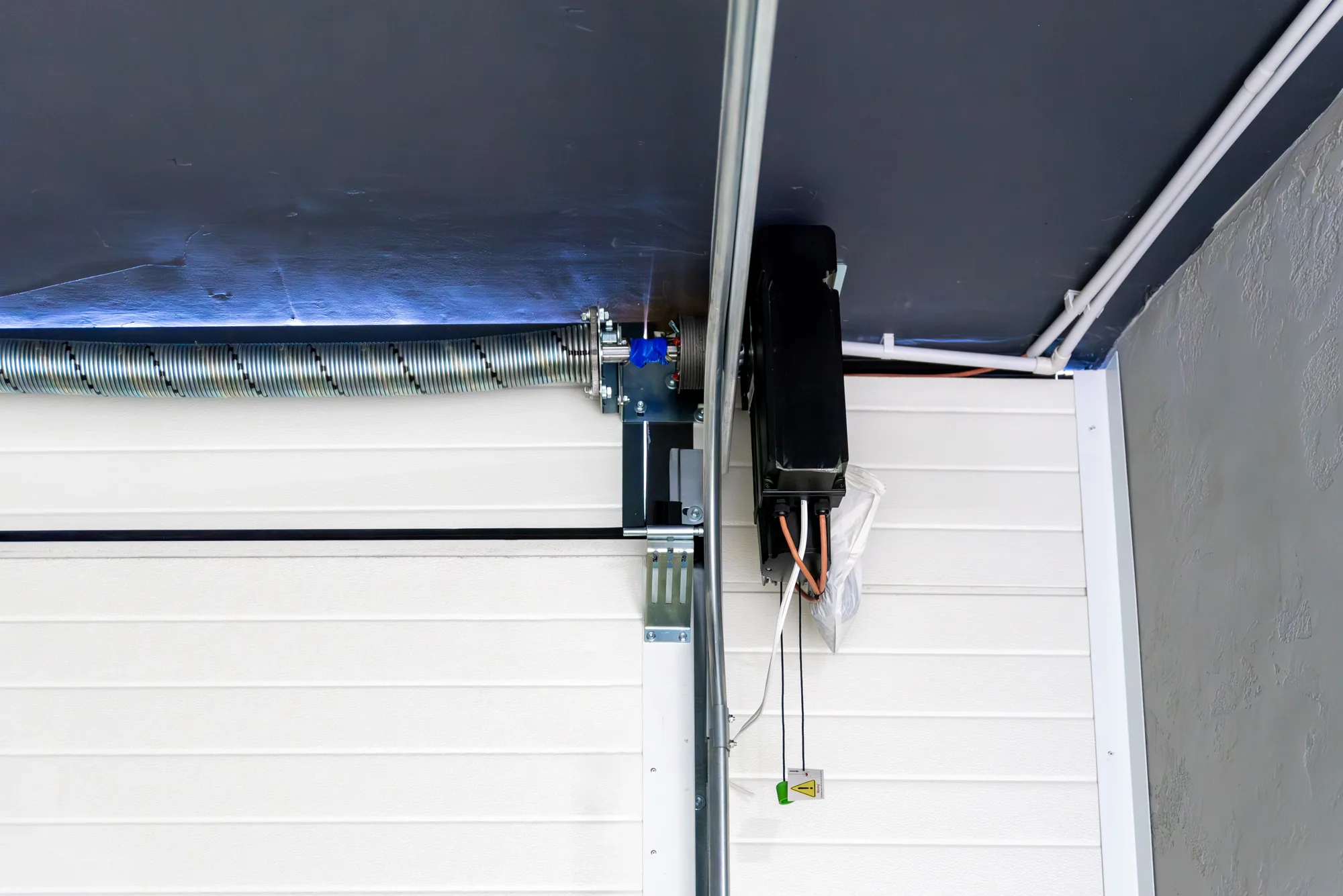 Vertical garage door track running down beside a white sectional door, with the torsion spring on its shaft and a side-mounted opener above