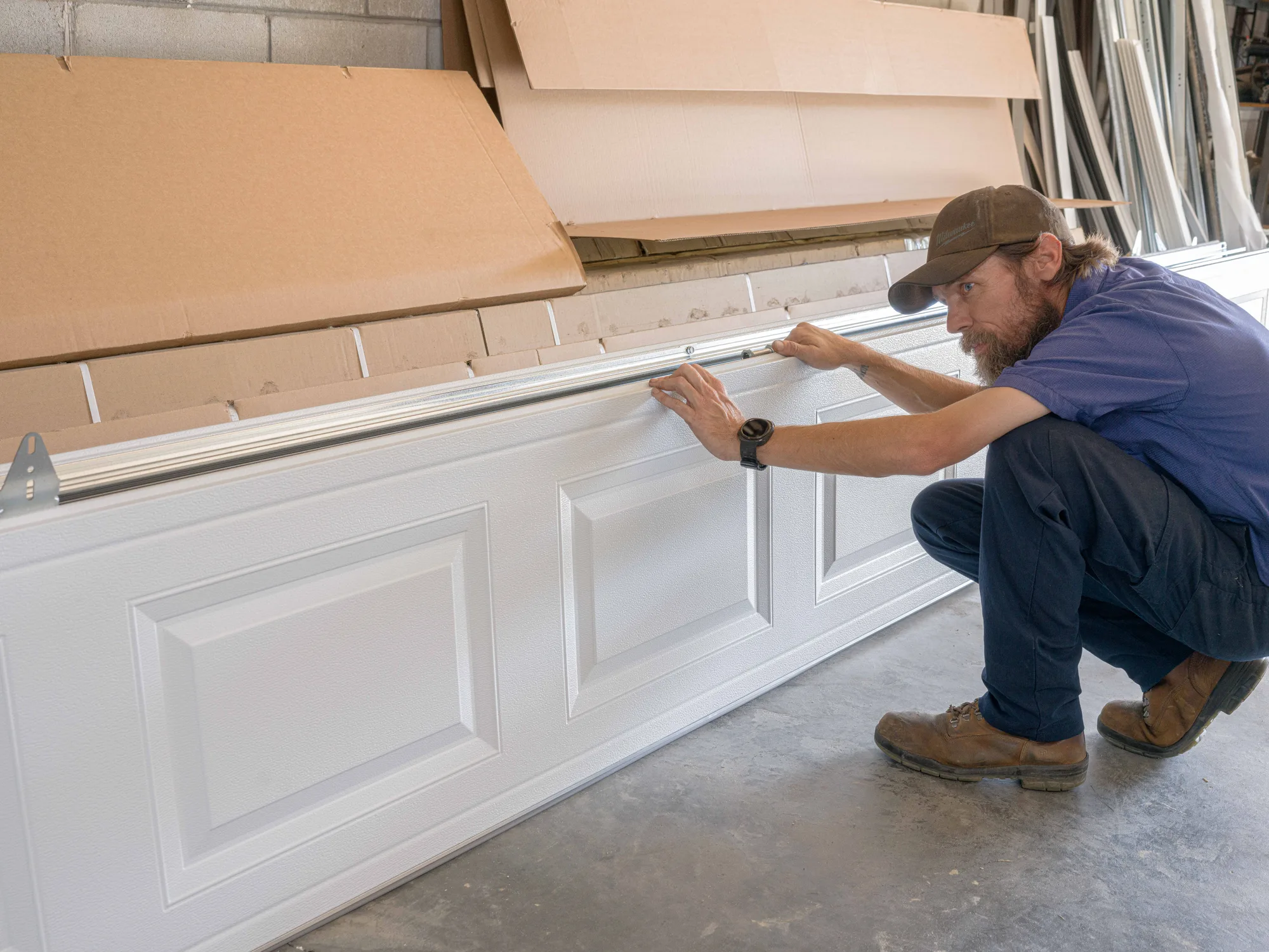 Technician in a Steve's Garage Doors work shirt fitting a strut along the top of a white raised-panel door section in the shop