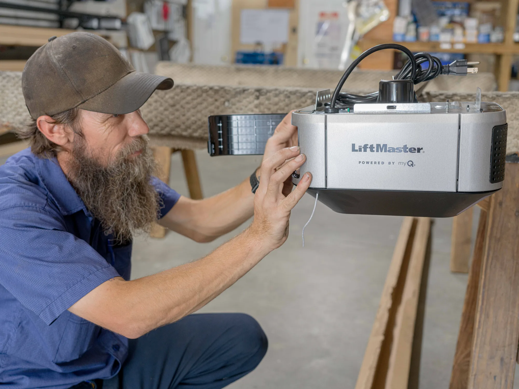 Technician in a blue work shirt and cap holding a LiftMaster opener head steady on a shop bench